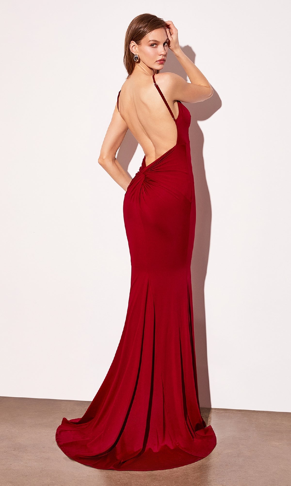 A woman in the deep red Astee T905 Open-Back Long Bodycon Prom Dress stands against a white wall, glancing over her shoulder.